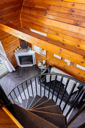 A spiral staircase leading to a wooden ceiling room with a fireplace, a sofa, and a small table with candles. - Starlit Hot Tub & Fire Pit | Modern Cabin for Two (Logan)