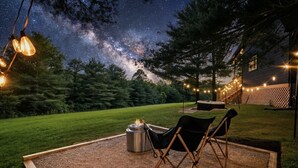 Een terras met vuurkorf, stoelen en lichtslingers onder een sterrenhemel.