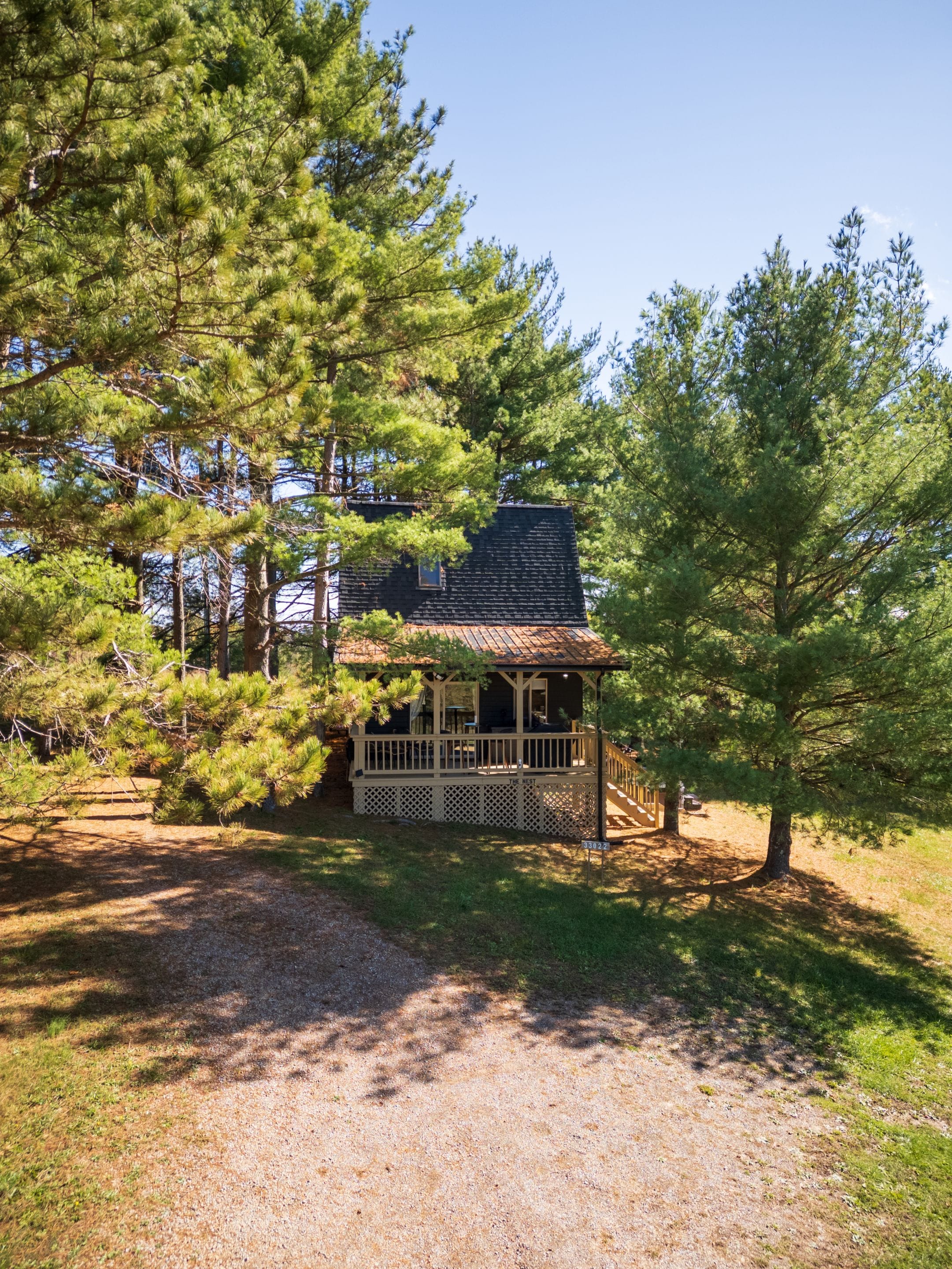 Une cabane dotée d’une véranda, entourée de pins et bordée d’un chemin de gravier.