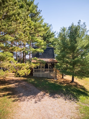 Une cabane dotée d’une véranda, entourée de pins et bordée d’un chemin de gravier.