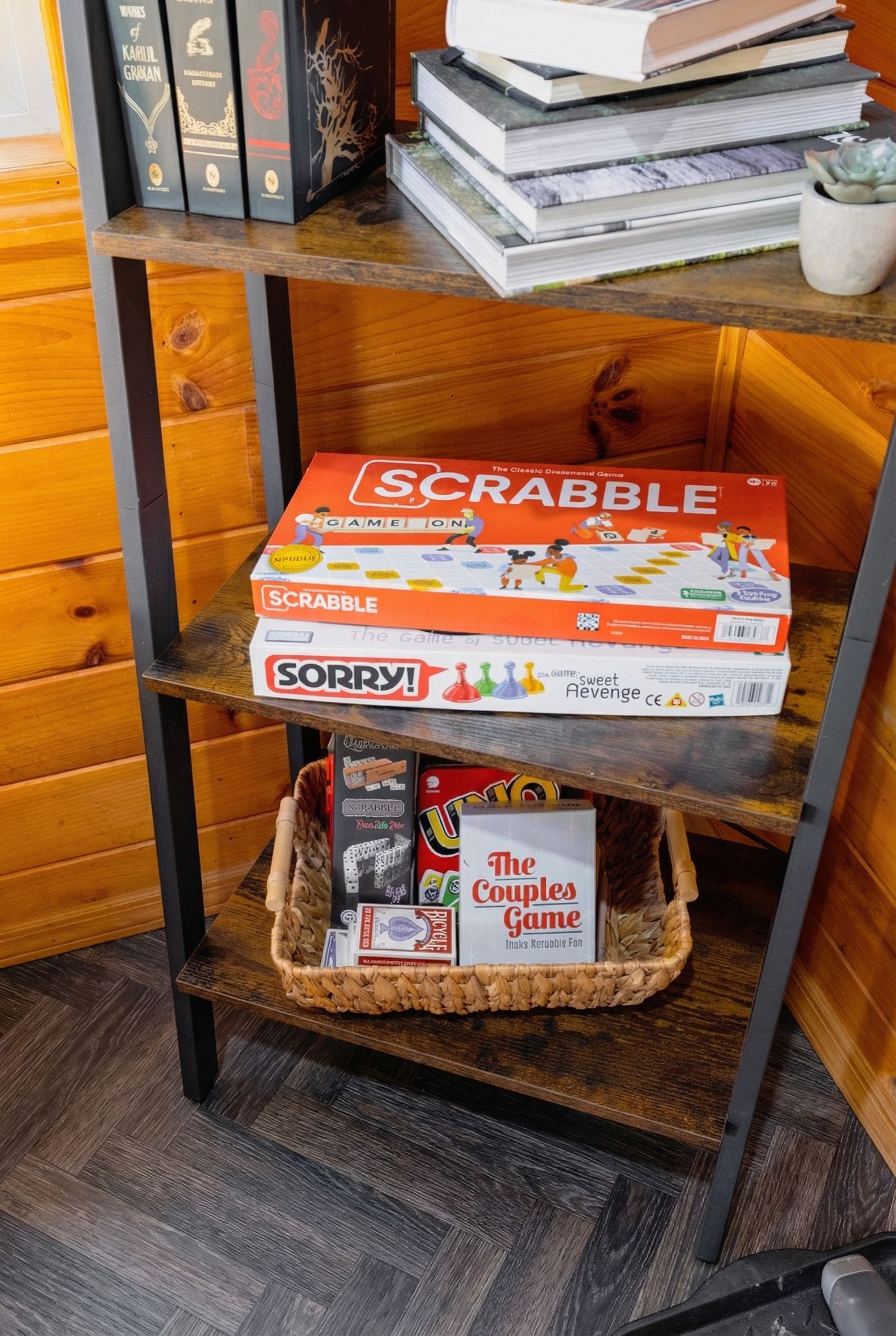 Una estantería de madera con libros, una caja de Scrabble, un juego de Sorry!, y una cesta con un juego para parejas.