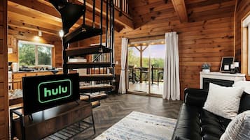 A wooden cabin interior with a living area, a fireplace, a dining area, and a large window.