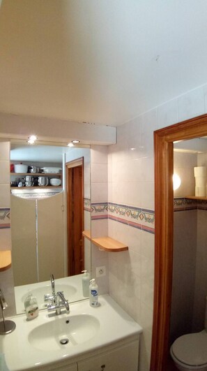 Une salle de bain avec un lavabo, un miroir et des toilettes. Une porte en bois donne accès à une cuisine.