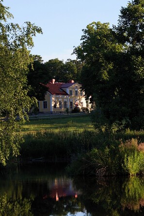 A large, two-story house with a red roof, surrounded by lush greenery and a body of water reflecting the structure.