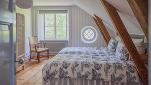 A bedroom with a sloped ceiling, a bed with a floral bedspread, a chair, a window with a view of greenery, and a radiator. - A grand, newly renovated villa in a castle setting (Stora Sundby)