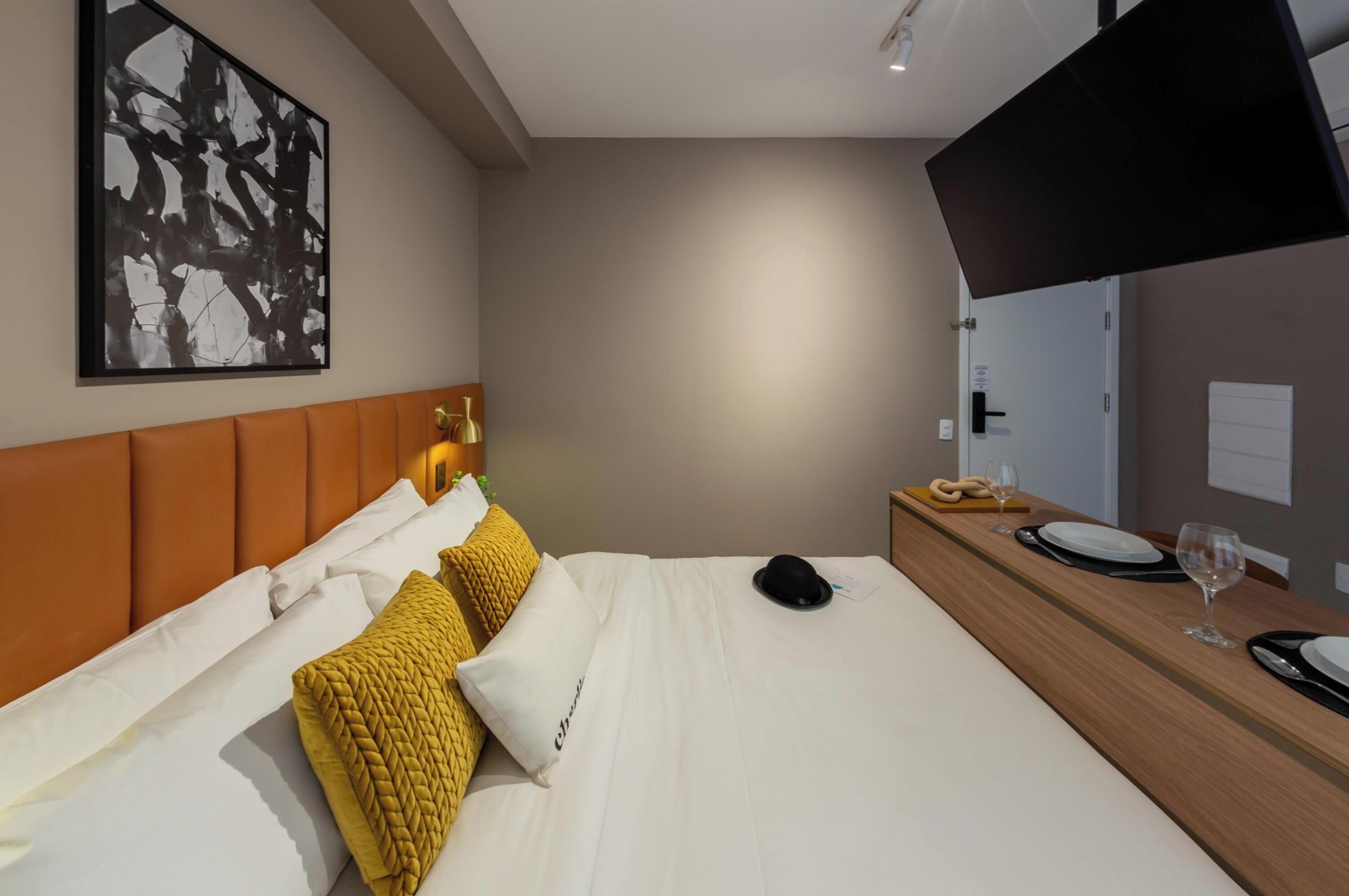 A modern hotel room with a large bed, a headboard, a bedside table with a mirror, and a wall-mounted artwork.