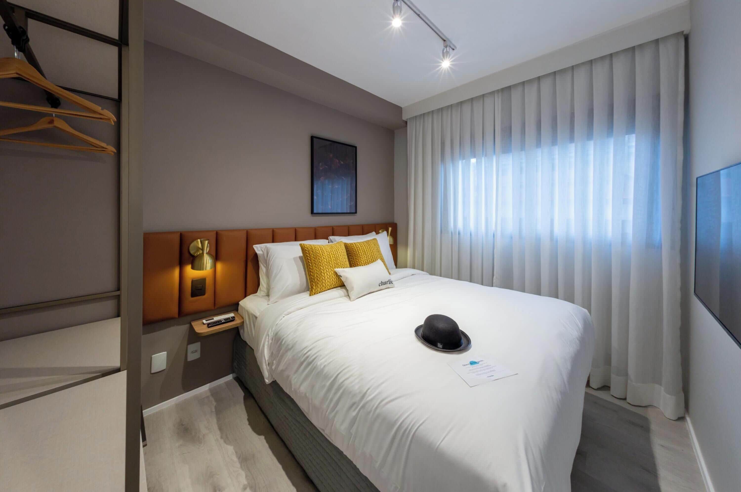 A modern hotel room with a large bed, a bedside table, a wall-mounted lamp, and a window with sheer curtains.