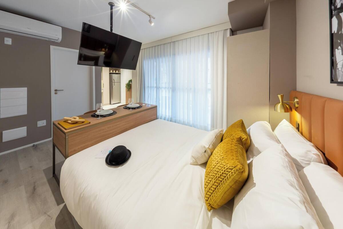 A modern hotel room with a large bed, a bedside table with a flat-screen TV, a desk with a chair, a window with curtains, and a wall-mounted light fixture.
