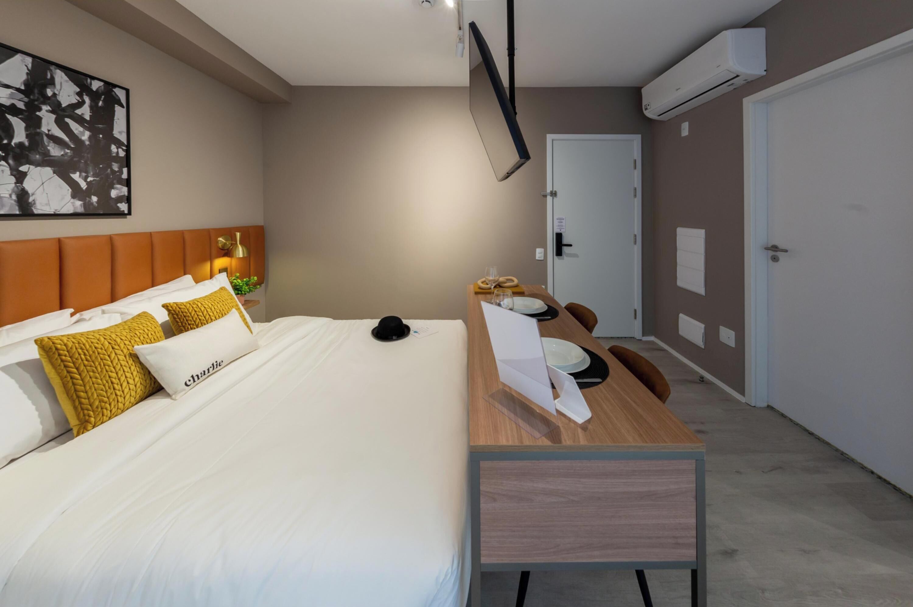 A hotel room with a bed, a desk, a chair, a bedside table, a wall-mounted TV, and an air conditioning unit.