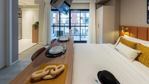 Une chambre d’hôtel comprenant un lit, une table à manger avec des chaises, un téléviseur fixé au mur et un balcon offrant une vue sur les bâtiments.