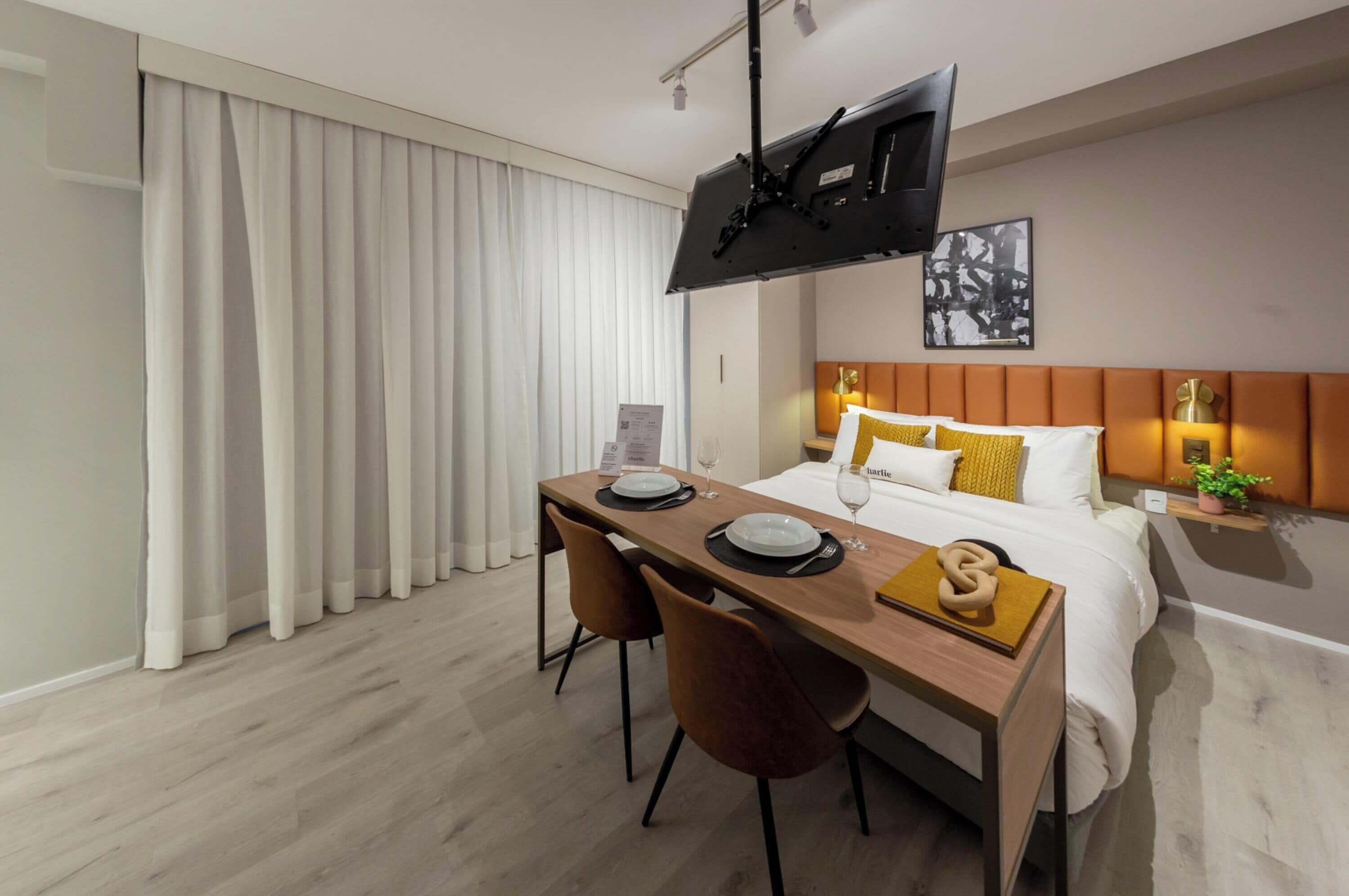 A hotel room with a bed, a TV mounted on the wall, a dining table with chairs, and a small side table with a plant.