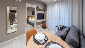 Une chambre d’hôtel moderne dotée d’un coin repas, d’un canapé, d’une télévision et d’une grande fenêtre avec des rideaux.
