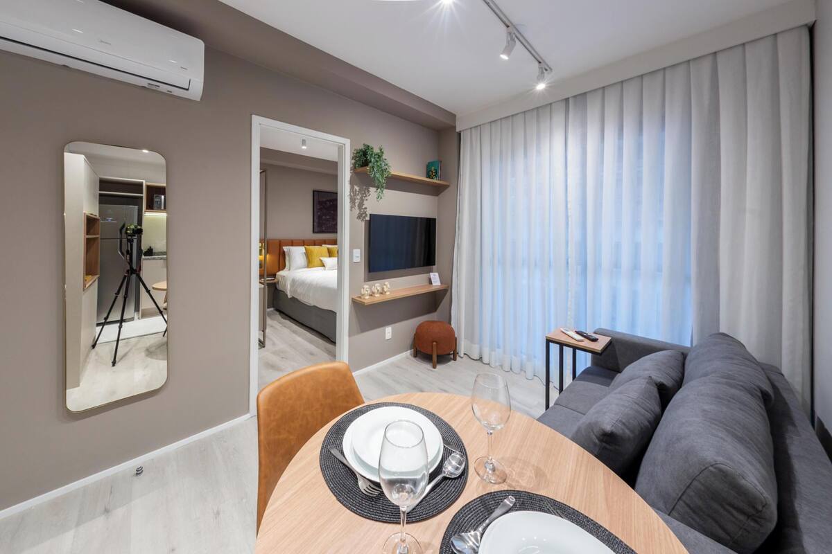 A modern hotel room with a dining area, a sofa, a TV, and a large window with curtains.