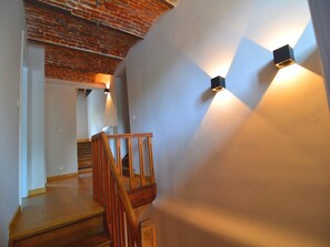 Un escalier avec des rampes en bois et un plafond en briques. Deux appliques murales sont fixées de part et d’autre de l’escalier.