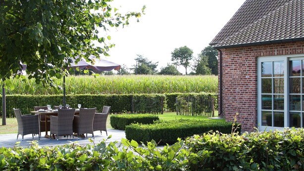An outdoor seating area with wicker chairs and a table, surrounded by well-maintained hedges and a brick building with large windows. - Peer Villa with Garden (Peer)