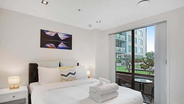 A hotel room with a large bed, a nightstand, a painting on the wall, and a balcony with a view of a building and greenery. - Quayside Quarters Apt close to Spark Arena (Auckland CBD)
