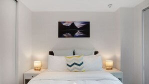 A neatly made bed with white linens and two bedside lamps. A painting of mountains and a lake hangs above the bed. - Quayside Quarters Apt close to Spark Arena (Auckland CBD)