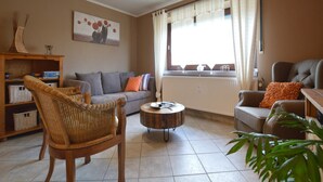 A living room with a grey sofa, a wicker chair, a wooden coffee table, and a window with curtains. - Waimes Retreat Near Forest (Waimes)