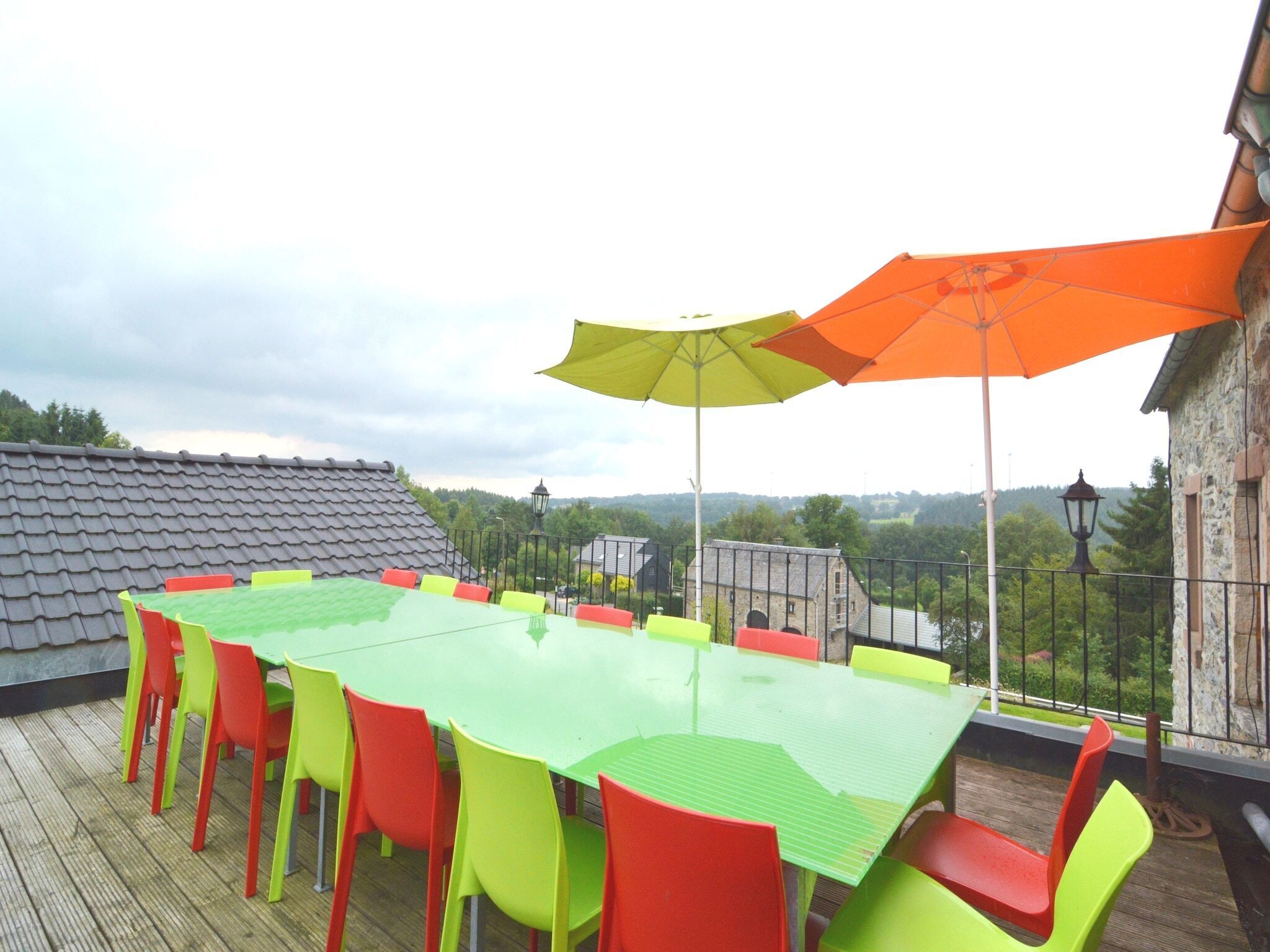 Una amplia mesa al aire libre con sillas de colores y dos sombrillas, ubicada en una terraza de madera con vistas a edificios y árboles.