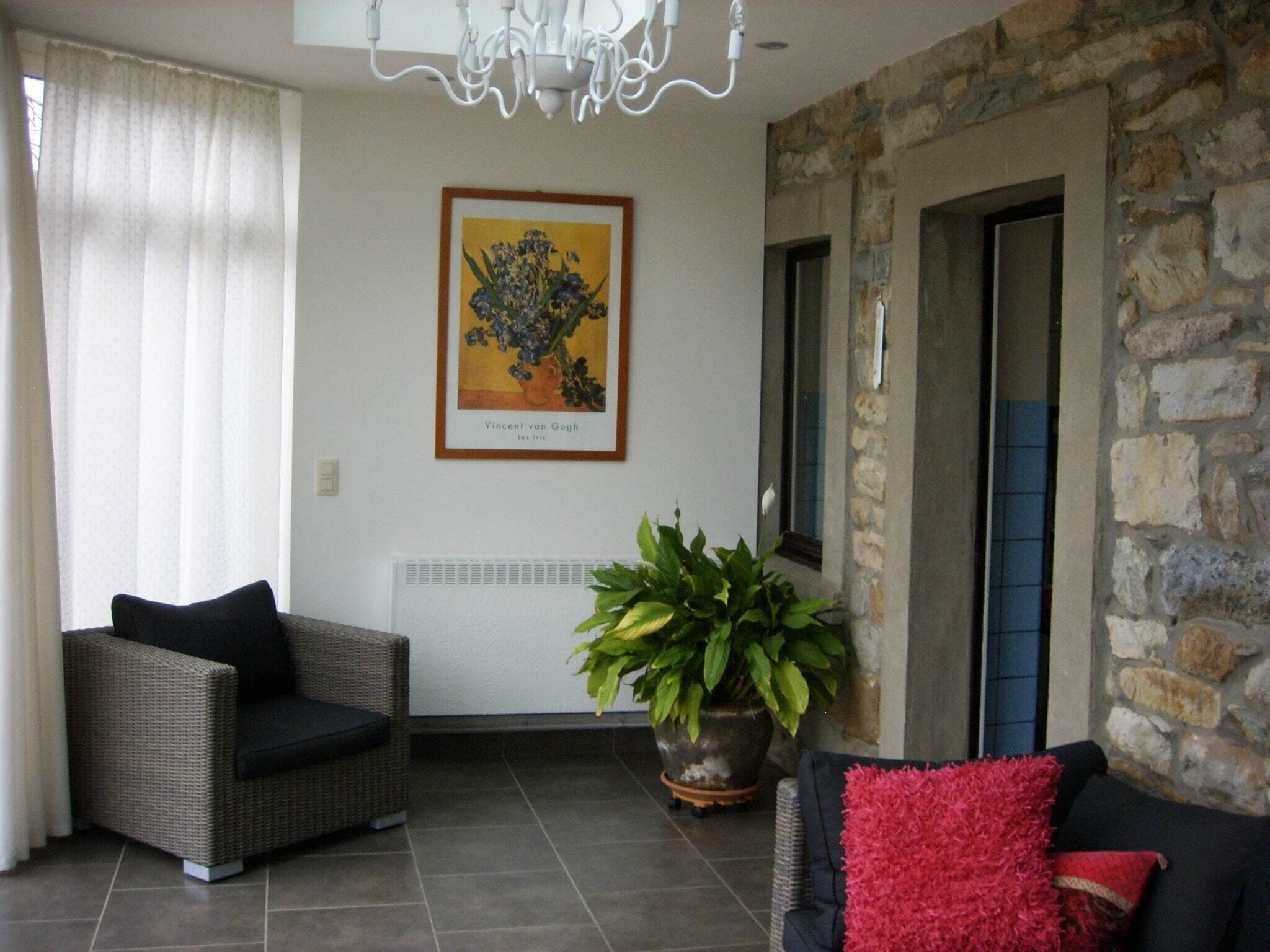 A living room with a wicker sofa, a red throw pillow, a potted plant, a framed artwork, and a stone wall.