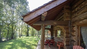 Una cabaña de troncos con terraza de madera, mobiliario exterior y vista del bosque circundante.