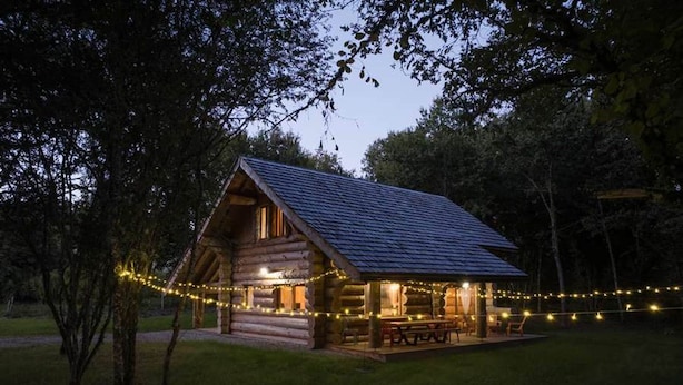 Una cabaña de madera con techo inclinado, rodeada de luces colgantes, y una mesa de picnic afuera.