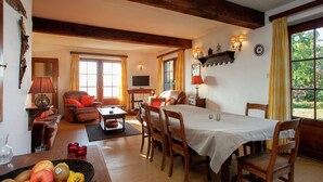 A spacious living room with a dining area, wooden beams, a television, and a large window with curtains.
