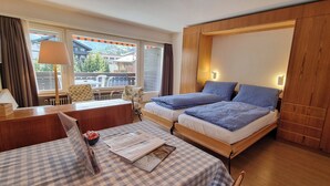 A hotel room with a checkered table, a bed with blue bedding, a wooden wardrobe, and a view of a mountain and buildings through large windows. - Haus Matten - Studio Findeln (Zermatt)