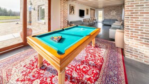 Une table de billard se trouve dans une pièce spacieuse, dotée d’un tapis rouge à motifs, d’un mur en briques et d’une grande fenêtre donnant sur un patio.