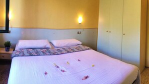 A neatly made bed with a floral bedspread and pillows, a bedside plant, and a wardrobe with a light-colored door. - Directly on the lake with terrace (Tre Capitelli)