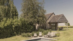 Un bâtiment rustique en pierre, avec un grand arbre devant, une piscine et un espace de détente extérieur.