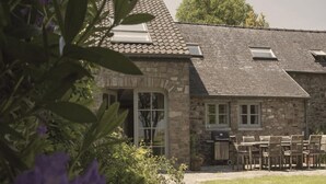 Un bâtiment en pierre avec un toit en tuiles, un patio couvert avec des meubles en bois et un jardin agrémenté de fleurs violettes.