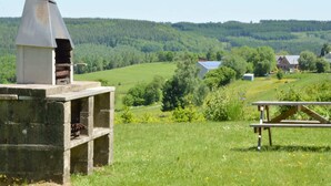 A rustic outdoor setting with a stone-built oven structure, a picnic table, and a view of a rural landscape with houses and trees. - Family Apartment; Bouillon and Semois River (Gros-Fays)
