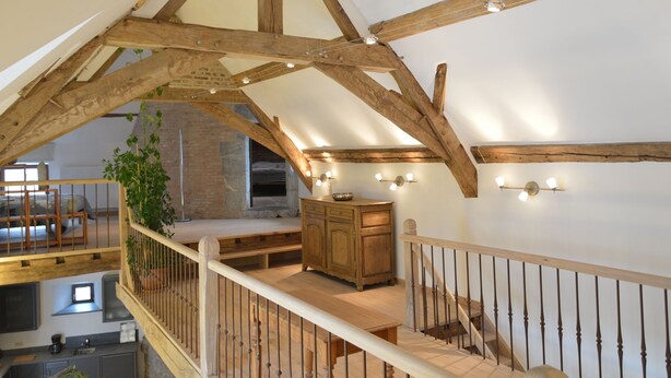 Ein Schlafzimmer im Loft-Stil mit freiliegenden Holzbalken, einer Holztreppe und Blick auf eine Küche darunter.