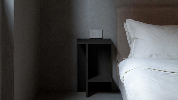 A minimalist hotel room with a single bed, a nightstand, and a small shelf. - Ho'c Suite (Milano)