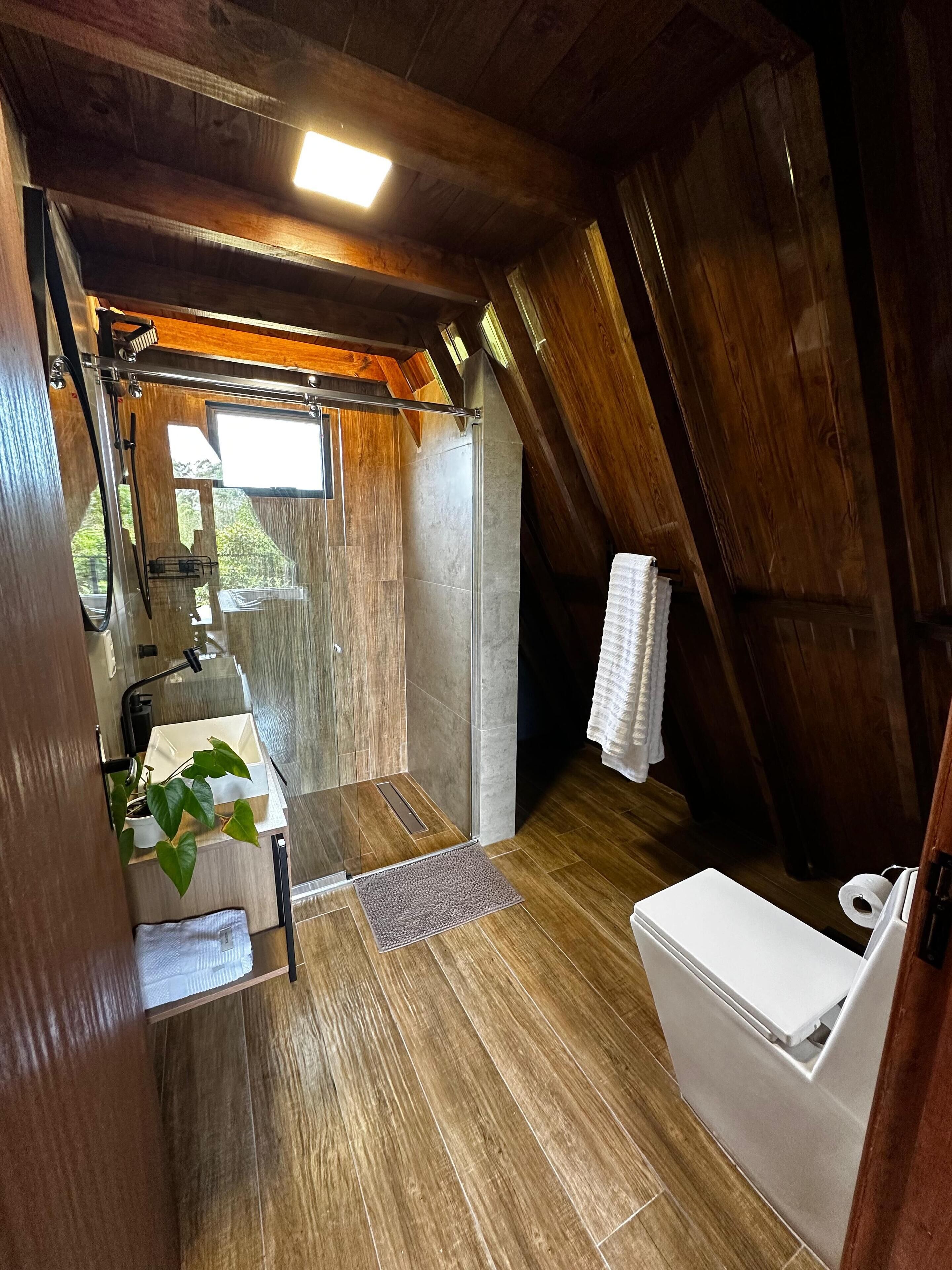 A compact, rustic bathroom with wooden beams, a shower, a toilet, and a sink with a plant.