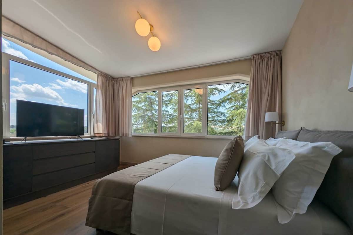 A bedroom with a large bed, a TV on a stand, and a window with curtains.