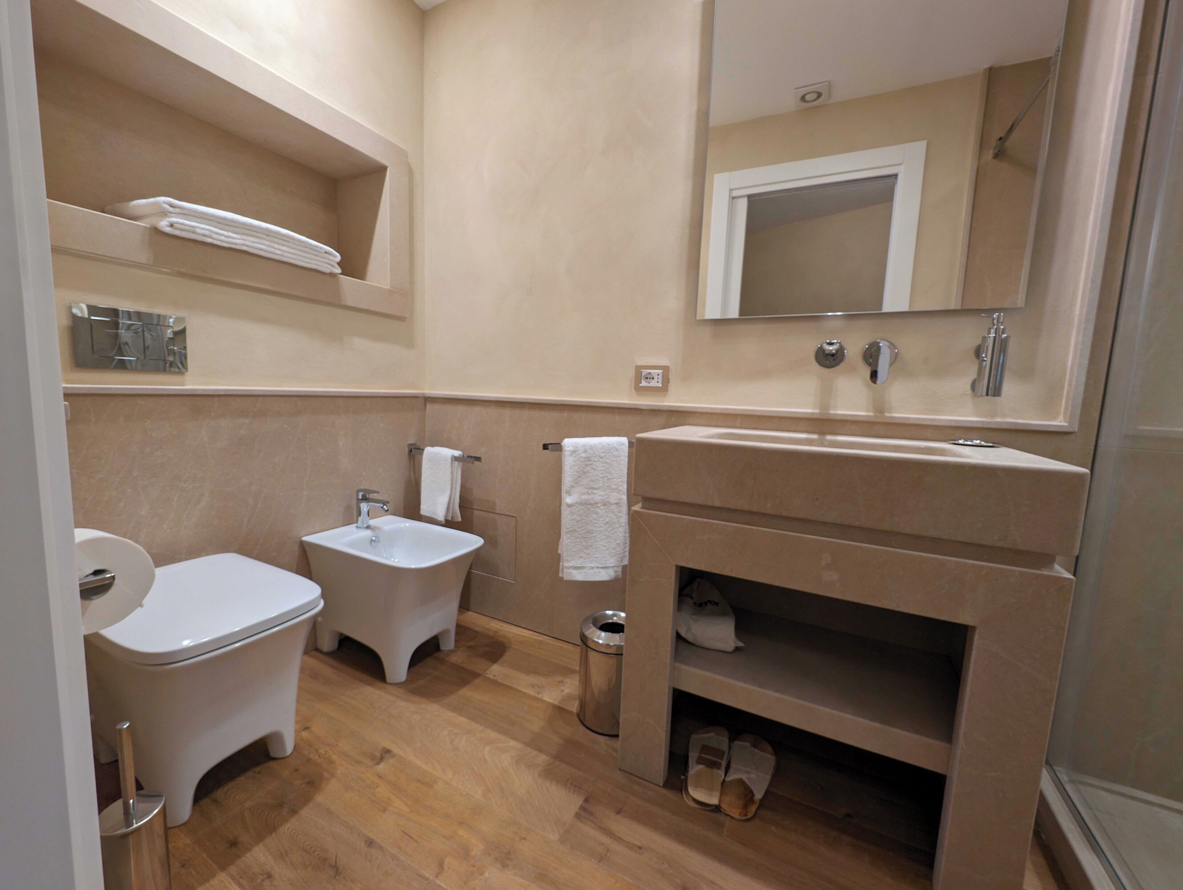 A compact bathroom with a toilet, bidet, sink, mirror, and a shelf with towels.