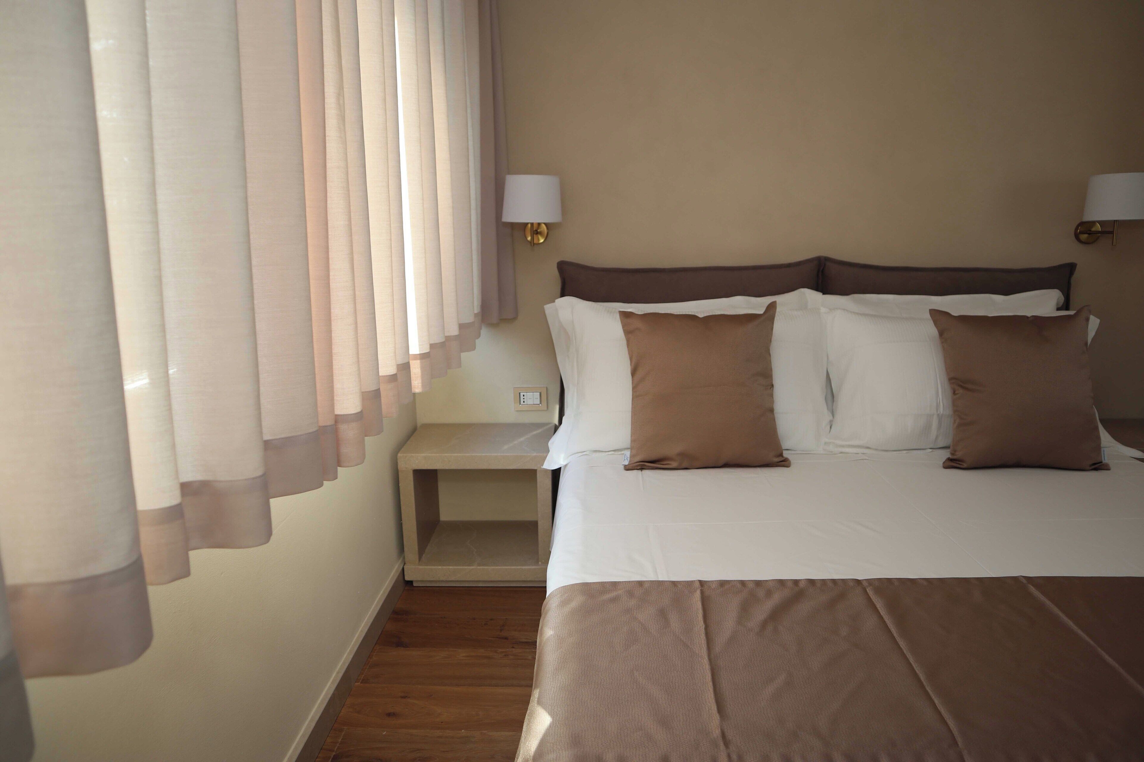 Una habitación de hotel con una cama grande, dos lámparas de noche, una mesita de noche y cortinas traslúcidas.