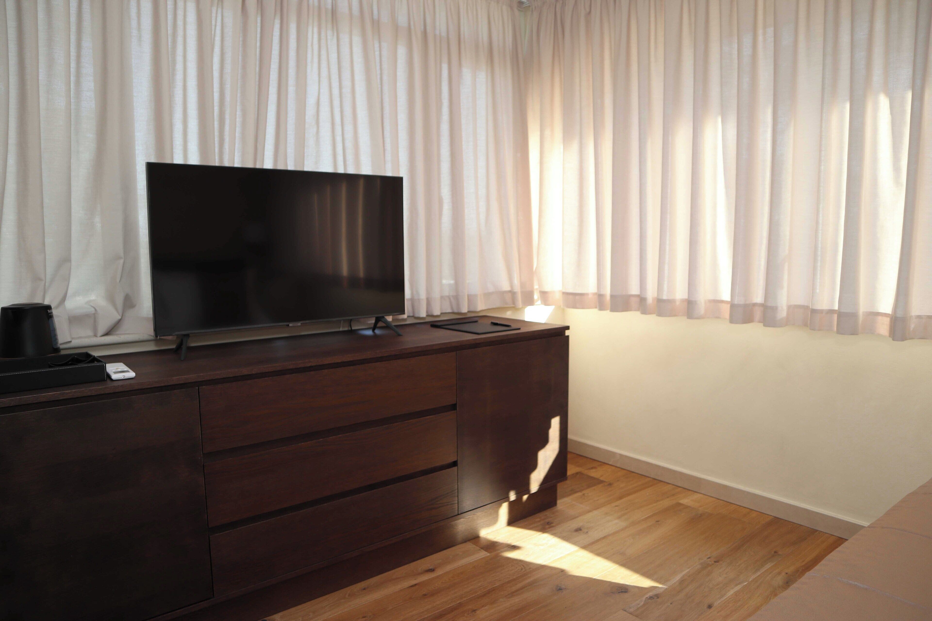 A room with a wooden dresser, a flat-screen TV, and sheer curtains.