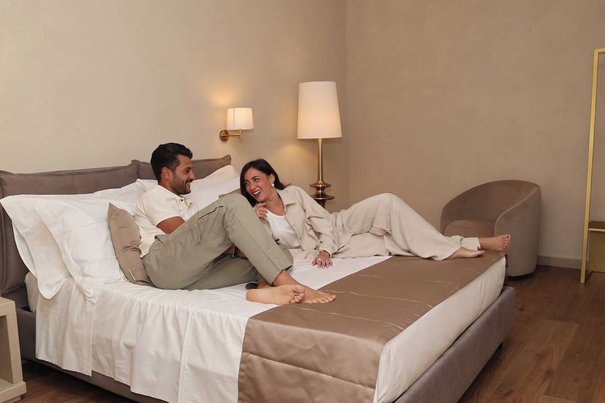 A bed with white linens and a brown accent, a bedside lamp, and a chair in a room with wooden flooring.