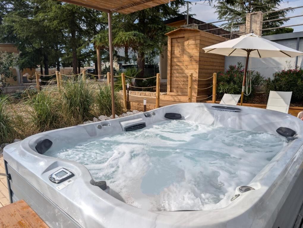 A hot tub with a wooden structure in the background, surrounded by a fenced area with chairs and an umbrella.