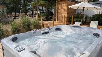A hot tub with a wooden structure in the background, surrounded by a fenced area with chairs and an umbrella.