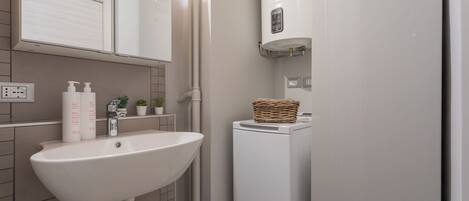 Une salle de bain avec un lavabo blanc, un miroir, un porte-serviettes, un lave-linge et une petite étagère contenant des articles de toilette.