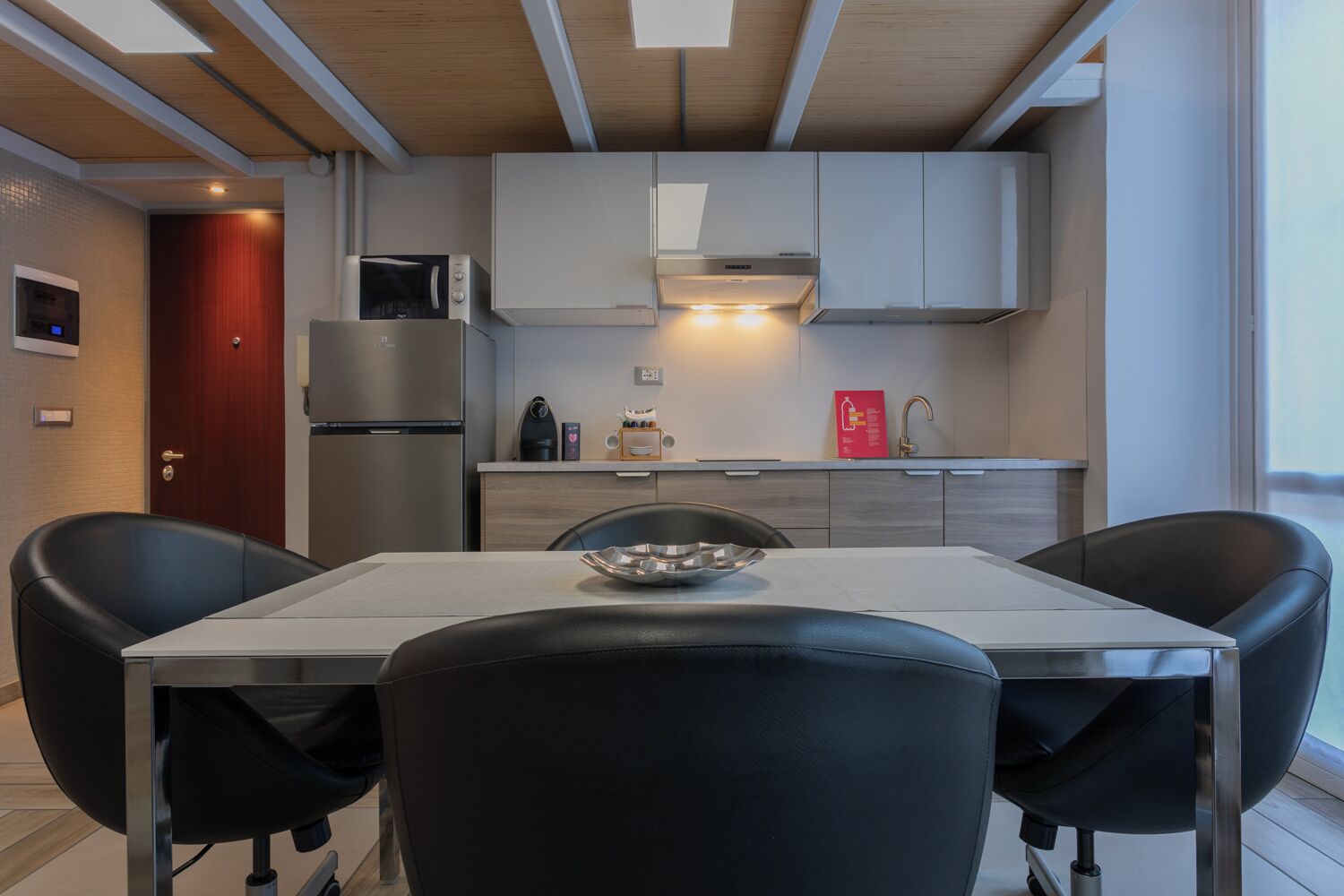 A modern kitchen with white cabinets, a stainless steel refrigerator, a microwave, and a gas stove. There is a dining table with two black chairs and a bowl on it.