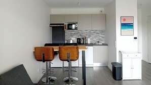 Eine moderne Küche mit weißen Schränken, einer schwarzen Arbeitsplatte und einer gefliesten Rückwand. Zwei hölzerne Barhocker und ein kleiner, weißer Schrank mit Schubladen sind ebenfalls vorhanden.