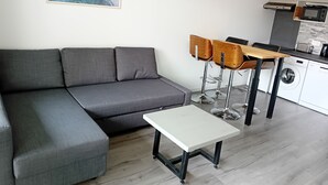 Ein modernes Wohnzimmer mit einem grauen Ecksofa, einem kleinen weißen Tisch und zwei Holzstühlen. Im Hintergrund ist eine Küchenzeile mit Waschmaschine und Mikrowelle erkennbar.