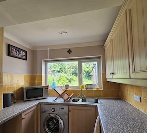 A compact kitchen with a washing machine, microwave, and a window with a view of greenery.