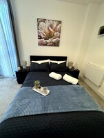 A bedroom with a large bed, a nightstand with a lamp, a painting on the wall, and a window with curtains.