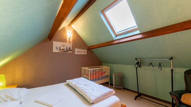 A bedroom with a sloped ceiling, a skylight, a crib, a radiator, a chair, and a bed with white linens. - Cozy Family Cottage in Érezée (Erezée)